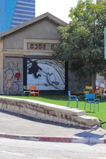 A small building with a graffiti-covered facade, featuring different artworks and abstract designs. In front of the building are several colorful chairs, arranged casually on artificial grass under a tree. The background shows a modern, blue glass building.