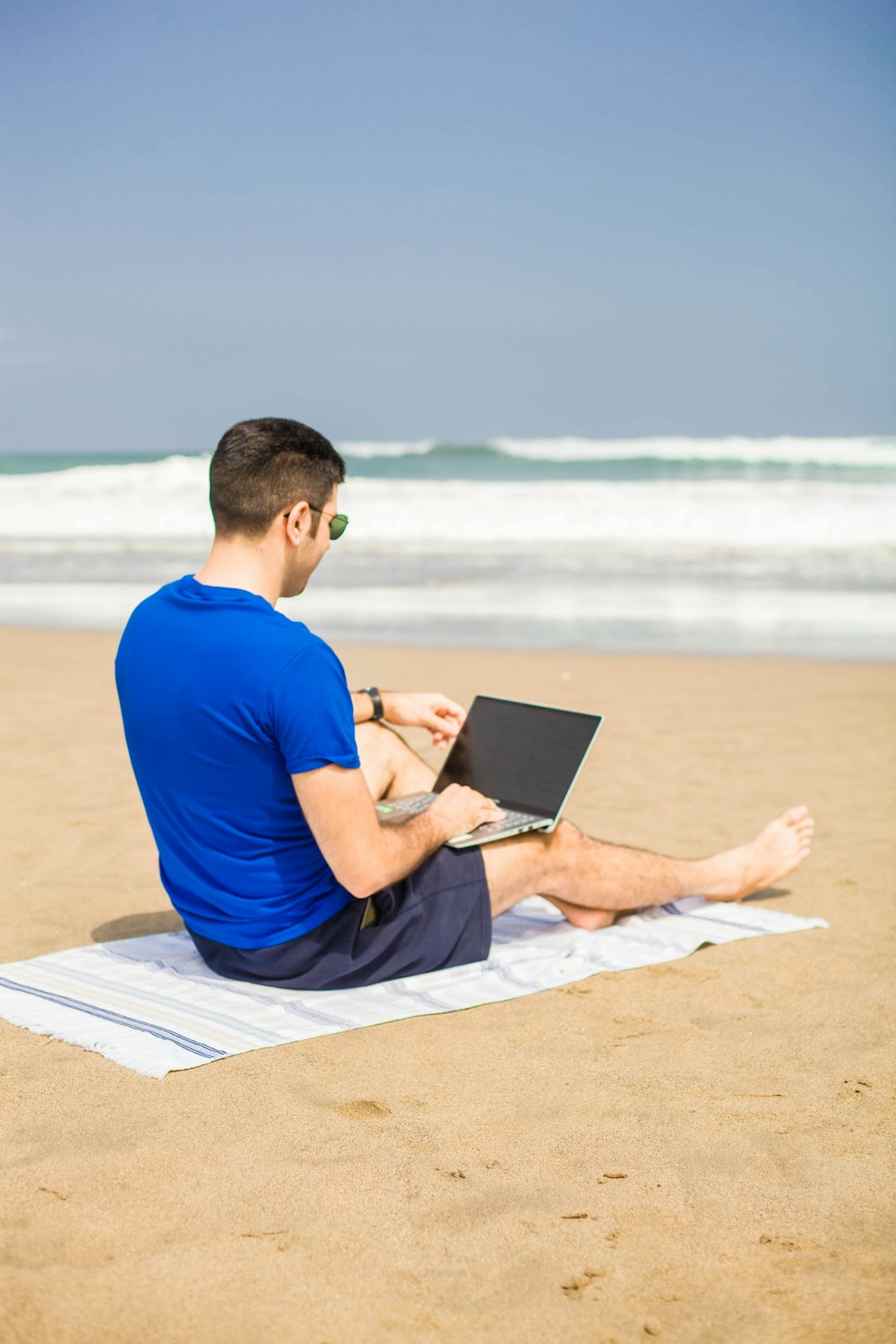 man in blue crew neck t-shirt sitting on white sand using black laptop computer during