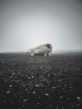 An abandoned, wrecked airplane lies on a desolate, barren landscape. The scene is enveloped in thick fog, enhancing the sense of isolation. The ground is strewn with small rocks and pebbles, and the horizon is barely visible.