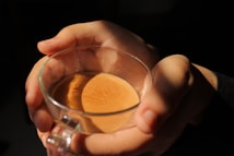 Hands are gently holding a glass cup filled with tea or coffee. Warm light illuminates the liquid, creating a cozy and inviting atmosphere.