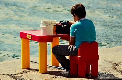 A young person wearing a teal shirt and dark pants sits on a small red chair at a colorful children's table. On the table, there is a white bucket and a black object. The scene is set outdoors beside a body of water.