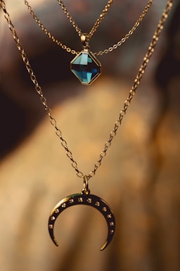A close-up of two necklaces made of gold chains. One necklace has a blue, hexagonal gemstone pendant, and the other has a golden crescent moon pendant with small gemstones embedded in it. The background is softly blurred with warm, earthy tones.