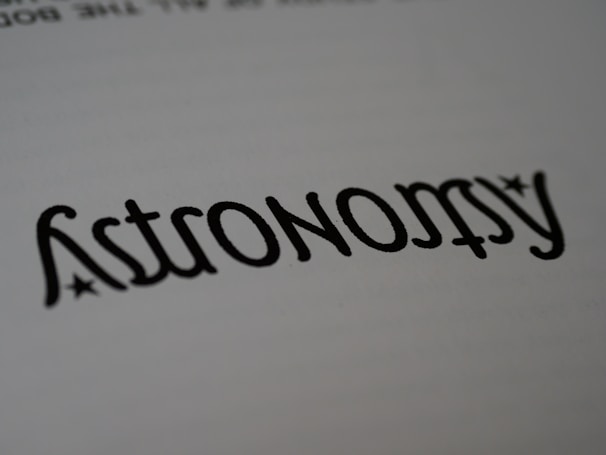 A close-up of the word 'Astronomy' written in a stylized font, resembling calligraphy. The text is black on a white background, with small star-like accents near the letters.