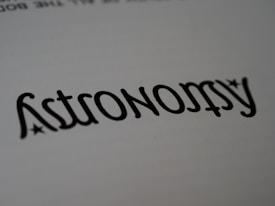 A close-up of the word 'Astronomy' written in a stylized font, resembling calligraphy. The text is black on a white background, with small star-like accents near the letters.