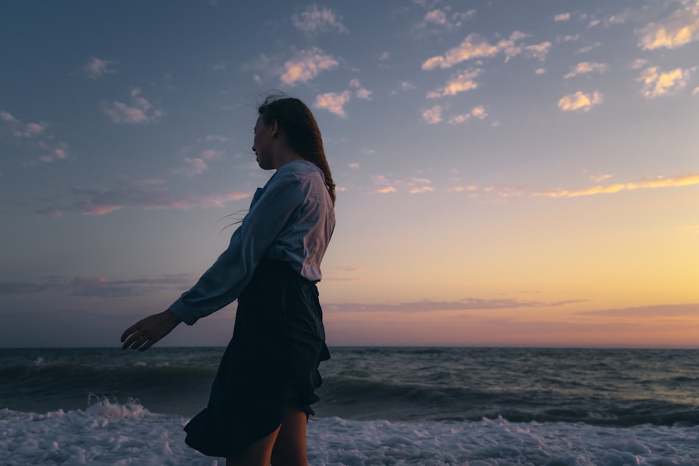 woman in white long sleeve shirt and black skirt standing on seashore during sunset