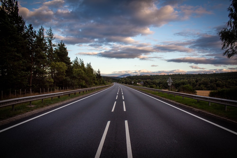 gray concrete road between green trees under white clouds and blue sky during daytime