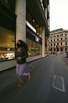 A person is jogging on the sidewalk in front of a pharmacy identified by a prominent sign. The building is modern with large windows and smooth columns. In the background is an older, more ornate building with multiple windows and elaborate architectural details.