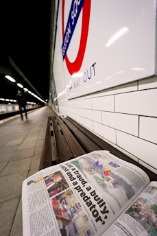 A bench located in an underground station with a newspaper resting on it, displaying a prominent headline. The station features classic white tiled walls and a red and blue roundel indicating 'Euston Square' with a 'way out' sign. The platform stretches into the distance under bright fluorescent lights, with a person walking in the background.