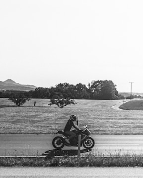 A black and white photograph of a person riding a motorcycle along a rural road. The landscape features open fields with a few scattered trees and a distant view of hills.