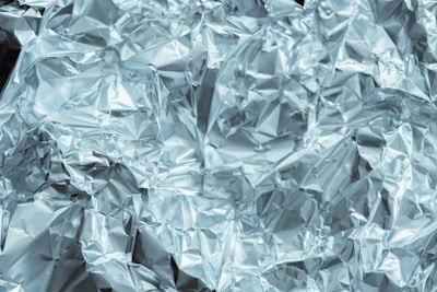 Crumpled and shiny silver material, likely aluminum foil, with various light reflections and creases creating an abstract and textured pattern.