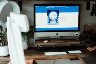 A modern workspace featuring a large iMac on a wooden desk, accompanied by a wireless keyboard and trackpad. The computer screen displays a website with a predominantly blue and white layout. Surrounding the setup are potted plants and books, adding a decorative touch to the setting. The chair in the foreground is white and ergonomically designed.