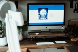 A modern workspace featuring a large iMac on a wooden desk, accompanied by a wireless keyboard and trackpad. The computer screen displays a website with a predominantly blue and white layout. Surrounding the setup are potted plants and books, adding a decorative touch to the setting. The chair in the foreground is white and ergonomically designed.