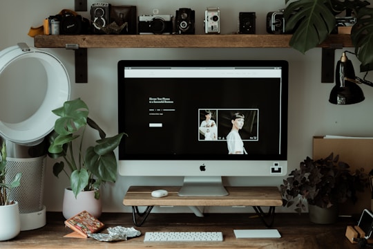 A home office setup features an iMac on a wooden desk surrounded by potted plants. The computer screen displays a minimalistic webpage with photography content. On the desk, various decorative items include a smartphone and a keyboard and mouse. Above the desk, a shelf holds vintage cameras. The desk lamp and air purifier add to the workspace functionality.