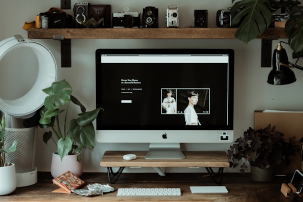 A home office setup features an iMac on a wooden desk surrounded by potted plants. The computer screen displays a minimalistic webpage with photography content. On the desk, various decorative items include a smartphone and a keyboard and mouse. Above the desk, a shelf holds vintage cameras. The desk lamp and air purifier add to the workspace functionality.
