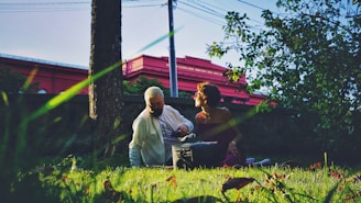 An elderly person and a younger individual are sitting on the grass in a park-like setting. Sunlight filters through the trees, creating a dappled effect on the ground. A red building is visible in the background. The scene conveys a relaxed and serene atmosphere.