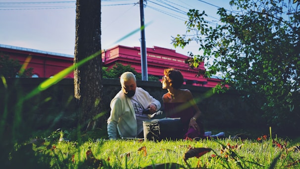 An elderly person and a younger individual are sitting on the grass in a park-like setting. Sunlight filters through the trees, creating a dappled effect on the ground. A red building is visible in the background. The scene conveys a relaxed and serene atmosphere.