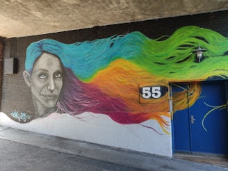 A vibrant mural depicting a woman's face with flowing hair in various colors. Her gaze is directed towards the viewer. The hair transitions from blue to green, yellow, orange, and pink. The number '55' is prominently displayed on a black sign on the wall. The artwork is situated on a dark brick wall under a concrete ceiling with a blue door nearby.