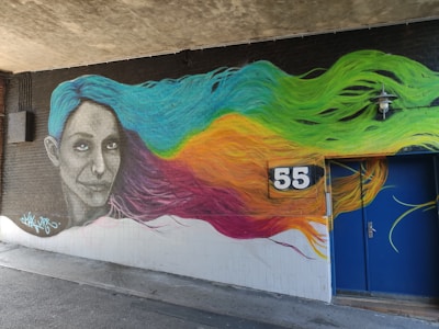 A vibrant mural depicting a woman's face with flowing hair in various colors. Her gaze is directed towards the viewer. The hair transitions from blue to green, yellow, orange, and pink. The number '55' is prominently displayed on a black sign on the wall. The artwork is situated on a dark brick wall under a concrete ceiling with a blue door nearby.