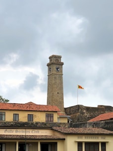 A tall, ancient stone clock tower stands prominently against a cloudy sky. Below, there is a building with a tiled roof displaying the words 'Galle Services Club Since 1847.' In the background, a wall of a fortress is visible with a flagpole flying a national flag.