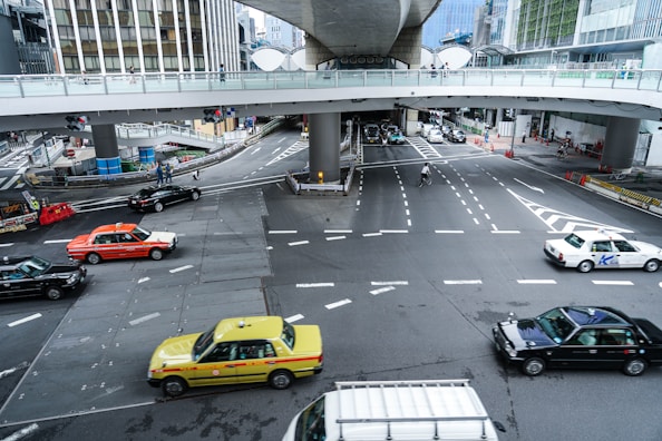 An urban intersection featuring multiple lanes of traffic beneath an overpass. Various cars of different colors, including a yellow taxi, a black car, and an orange and white taxi, are seen navigating the intersection. Pedestrians are visible on the overpass walkway, while cyclists traverse the road below. The area is surrounded by tall buildings and construction barriers.