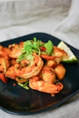 A plate of cooked shrimp seasoned with herbs and spices, garnished with fresh cilantro and a wedge of lime on the side. The dish is presented on a dark blue plate with a light fabric in the background.