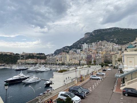 A coastal cityscape featuring a marina filled with luxury yachts in the foreground. Tall residential and commercial buildings rise against a backdrop of a large, rugged hillside. The sky is overcast with thick clouds, creating a moody atmosphere. Cars are parked along a road beside a building with classical architecture.