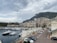 A coastal cityscape featuring a marina filled with luxury yachts in the foreground. Tall residential and commercial buildings rise against a backdrop of a large, rugged hillside. The sky is overcast with thick clouds, creating a moody atmosphere. Cars are parked along a road beside a building with classical architecture.
