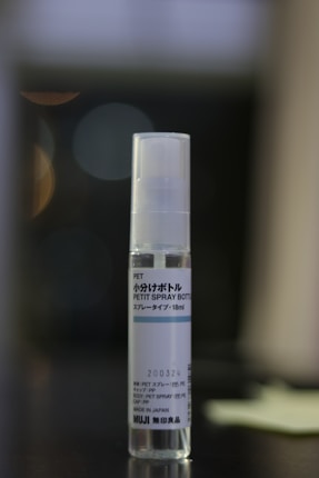 A transparent spray bottle labeled 'Petit Spray Bottle' from the brand Muji is centered against a blurred, dark background with soft lighting. Text in both Japanese and English is visible on the label.