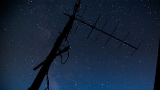 A silhouette of a large antenna against a starry night sky. The stars are numerous and scattered across a dark blue to black gradient, creating a serene and expansive atmosphere.