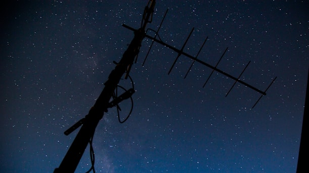 A silhouette of a large antenna against a starry night sky. The stars are numerous and scattered across a dark blue to black gradient, creating a serene and expansive atmosphere.