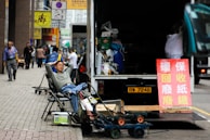 A man is reclining on a folding chair on a city sidewalk near an open truck loaded with various items. Nearby, a red sign with Chinese characters stands next to the truck. The street is bustling with a few pedestrians walking past high-rise buildings, and various carts and other miscellaneous items are scattered around.