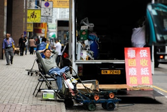 A man is reclining on a folding chair on a city sidewalk near an open truck loaded with various items. Nearby, a red sign with Chinese characters stands next to the truck. The street is bustling with a few pedestrians walking past high-rise buildings, and various carts and other miscellaneous items are scattered around.