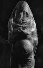 A pregnant person is wrapped in transparent plastic, creating a stark contrast against the dark background. The plastic tightly encases the figure, emphasizing the contours of their body.
