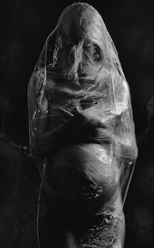 A pregnant person is wrapped in transparent plastic, creating a stark contrast against the dark background. The plastic tightly encases the figure, emphasizing the contours of their body.