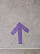 A purple arrow painted on a cracked concrete surface, pointing upwards. The texture of the concrete and the vivid color of the arrow create a striking contrast.