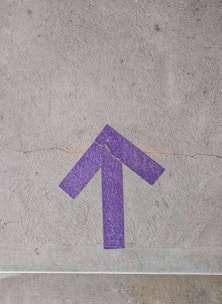 A purple arrow painted on a cracked concrete surface, pointing upwards. The texture of the concrete and the vivid color of the arrow create a striking contrast.