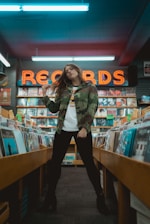 A person wearing a camouflage jacket and black pants stands in a record store, surrounded by racks filled with vinyl records. The neon sign in the background prominently displays the word 'RECORDS'. The setting has a retro and artistic vibe.