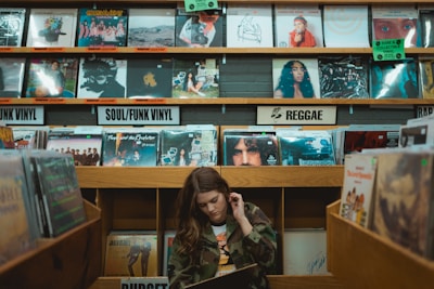 A person is browsing through vinyl records in a music store. The shelves are filled with a variety of record covers categorized by genres such as 'Soul/Funk Vinyl' and 'Reggae.' The ambiance is retro and nostalgic, with colorful album art visible on the shelves.
