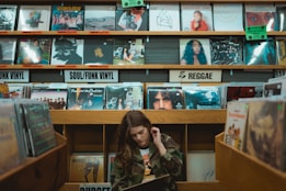 A person is browsing through vinyl records in a music store. The shelves are filled with a variety of record covers categorized by genres such as 'Soul/Funk Vinyl' and 'Reggae.' The ambiance is retro and nostalgic, with colorful album art visible on the shelves.