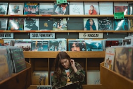 A person is browsing through vinyl records in a music store. The shelves are filled with a variety of record covers categorized by genres such as 'Soul/Funk Vinyl' and 'Reggae.' The ambiance is retro and nostalgic, with colorful album art visible on the shelves.