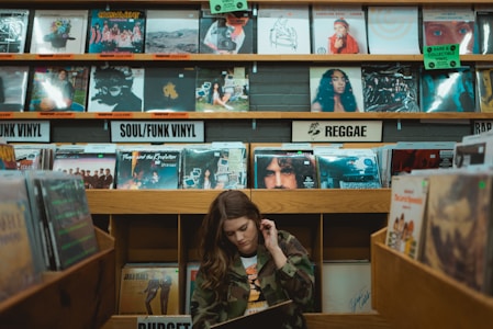 A person is browsing through vinyl records in a music store. The shelves are filled with a variety of record covers categorized by genres such as 'Soul/Funk Vinyl' and 'Reggae.' The ambiance is retro and nostalgic, with colorful album art visible on the shelves.