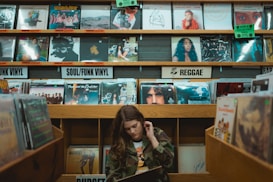 A person is browsing through vinyl records in a music store. The shelves are filled with a variety of record covers categorized by genres such as 'Soul/Funk Vinyl' and 'Reggae.' The ambiance is retro and nostalgic, with colorful album art visible on the shelves.