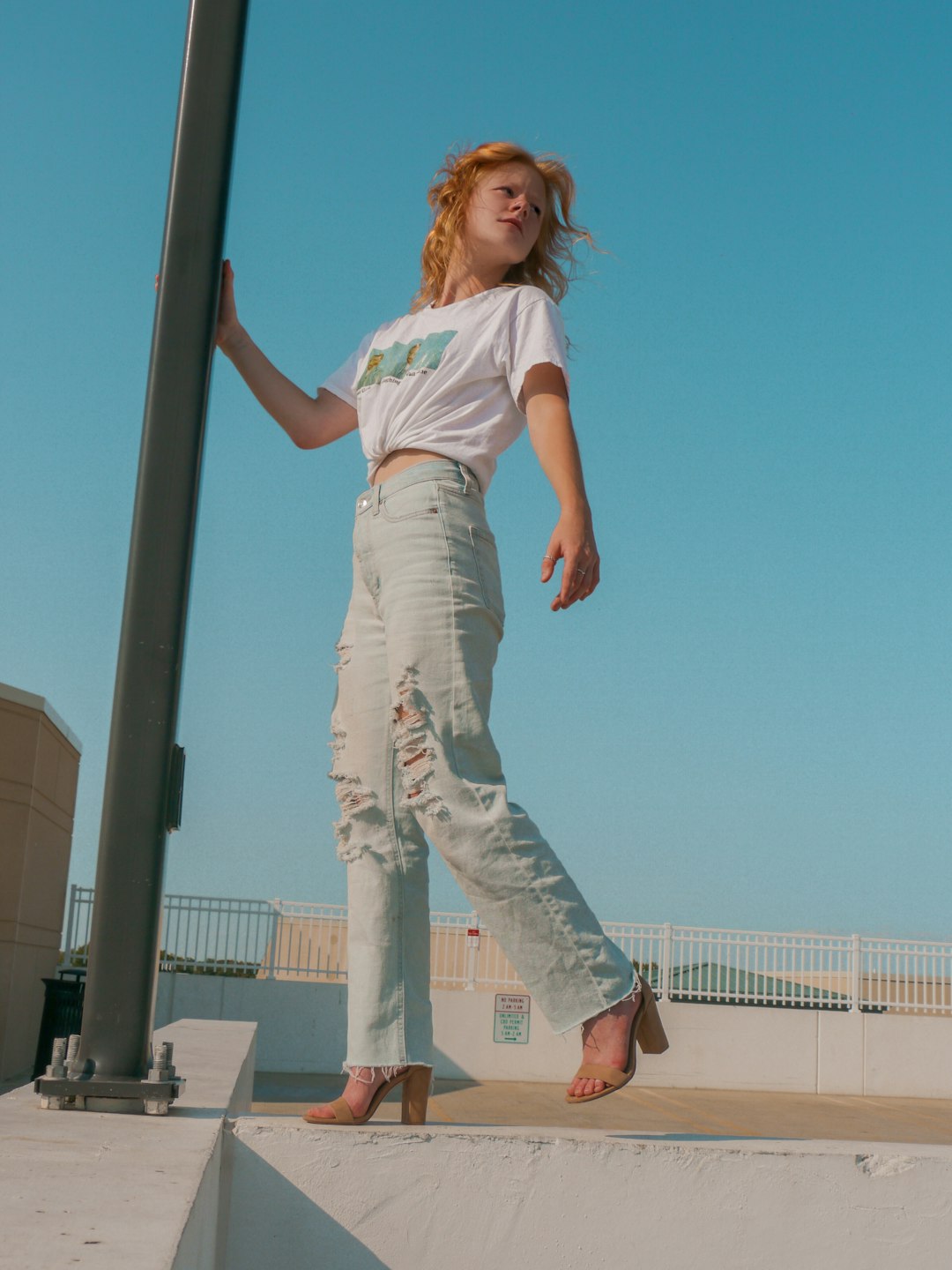 girl in white t-shirt and blue denim jeans standing on gray metal pole during daytime