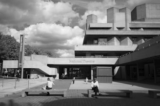 A black and white image of a modern, Brutalist-style building labeled 'National Theatre.' The structure is expansive with a blocky, geometric design. In the foreground, people sit on benches in an open plaza area, while others walk around. A large sign on the left advertises a seasonal event. Fluffy clouds fill the partly sunny sky, casting contrasting shadows on the building facades.