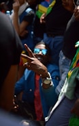 A group of people gathered together with one person in the foreground making a peace sign with their hand. This individual is wearing sunglasses and a blue jacket, creating a focal point with a lively and engaging presence.