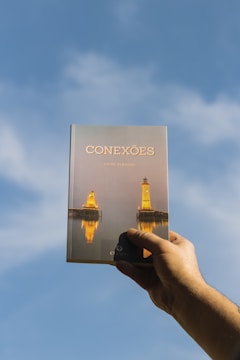 A hand is holding a book titled 'Conexões' against a clear blue sky. The cover features an image of two lighthouses reflected in water, illuminated in a warm, golden light.