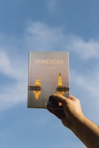 A hand is holding a book titled 'Conexões' against a clear blue sky. The cover features an image of two lighthouses reflected in water, illuminated in a warm, golden light.