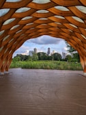 An architectural wooden structure frames a view of a green park with several skyscrapers in the background under a partly cloudy sky. The structure has an intricate, honeycomb pattern and is composed of curved wooden beams.