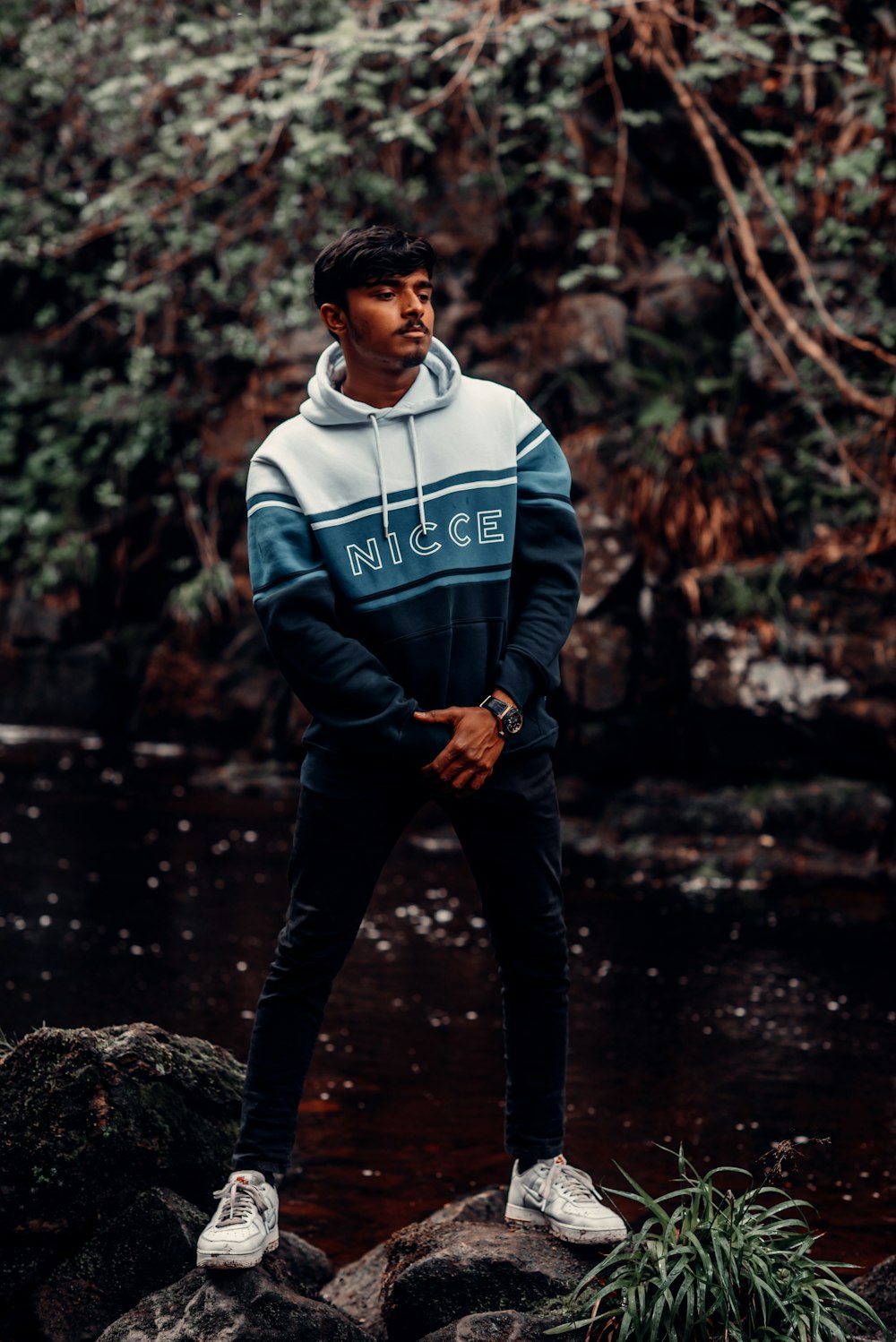 man in blue and white adidas hoodie and black pants standing on rock in water