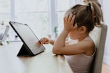 A young child with their hair in a ponytail sits at a table, interacting with a tablet device. The child rests one hand against their face while using their other hand to touch the screen. Natural light filters through large windows in the background, creating a soft and calm environment.